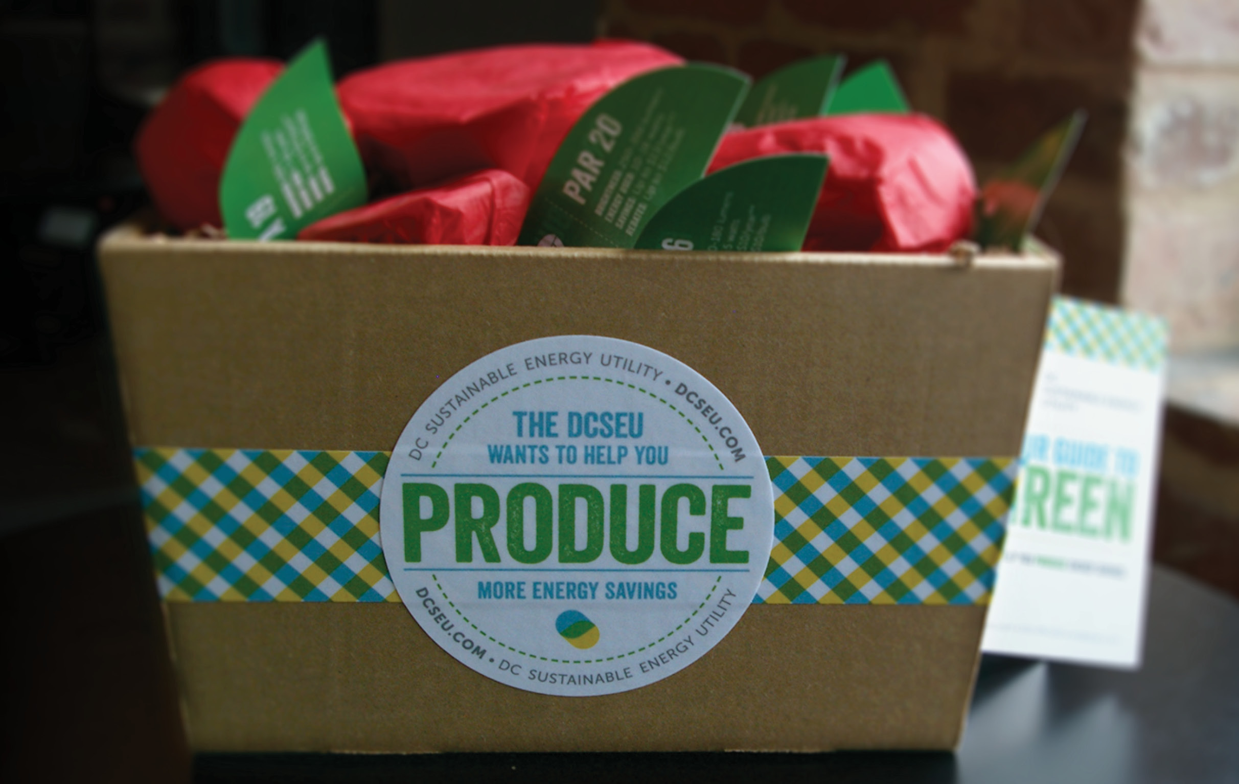 A white pick-your-own produce box filled with light bulbs wrapped in red tissue paper to look like apples. Green paper leaves identify which bulb is wrapping in each individual package. A hanging boolet is attached to the box by twine; it reads Your Guide to Green.