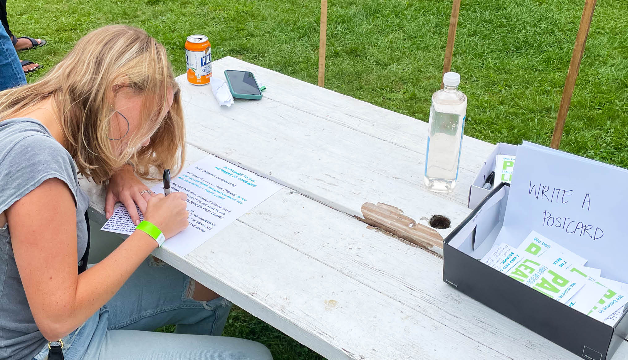 A person sitting at a picnic table, writing a postcard to thier congress member